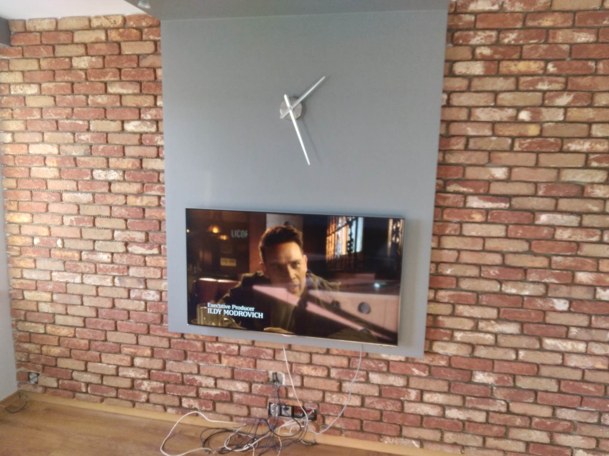 Ściana z cegły dekoracyjnej w salonie w Krakowie, z zamontowanym telewizorem i zegarem. Widoczne kable. Wykończenie wnętrza w stylu industrialnym.