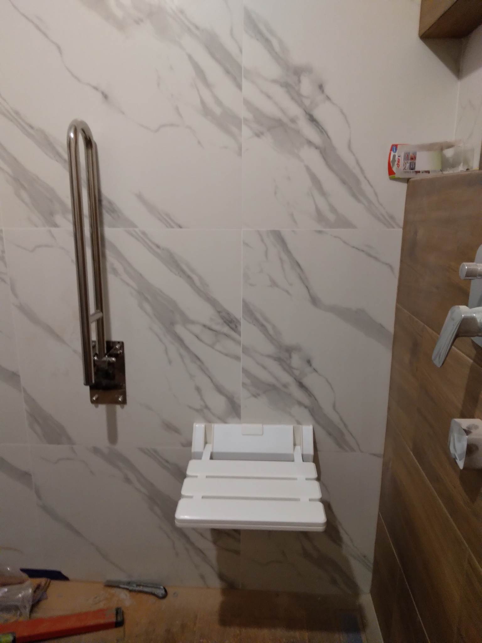 Biała, składana ławeczka prysznicowa i uchwyt zamontowane na ścianie z płytek imitujących marmur. Widoczne narzędzia na blacie sugerują trwający montaż.