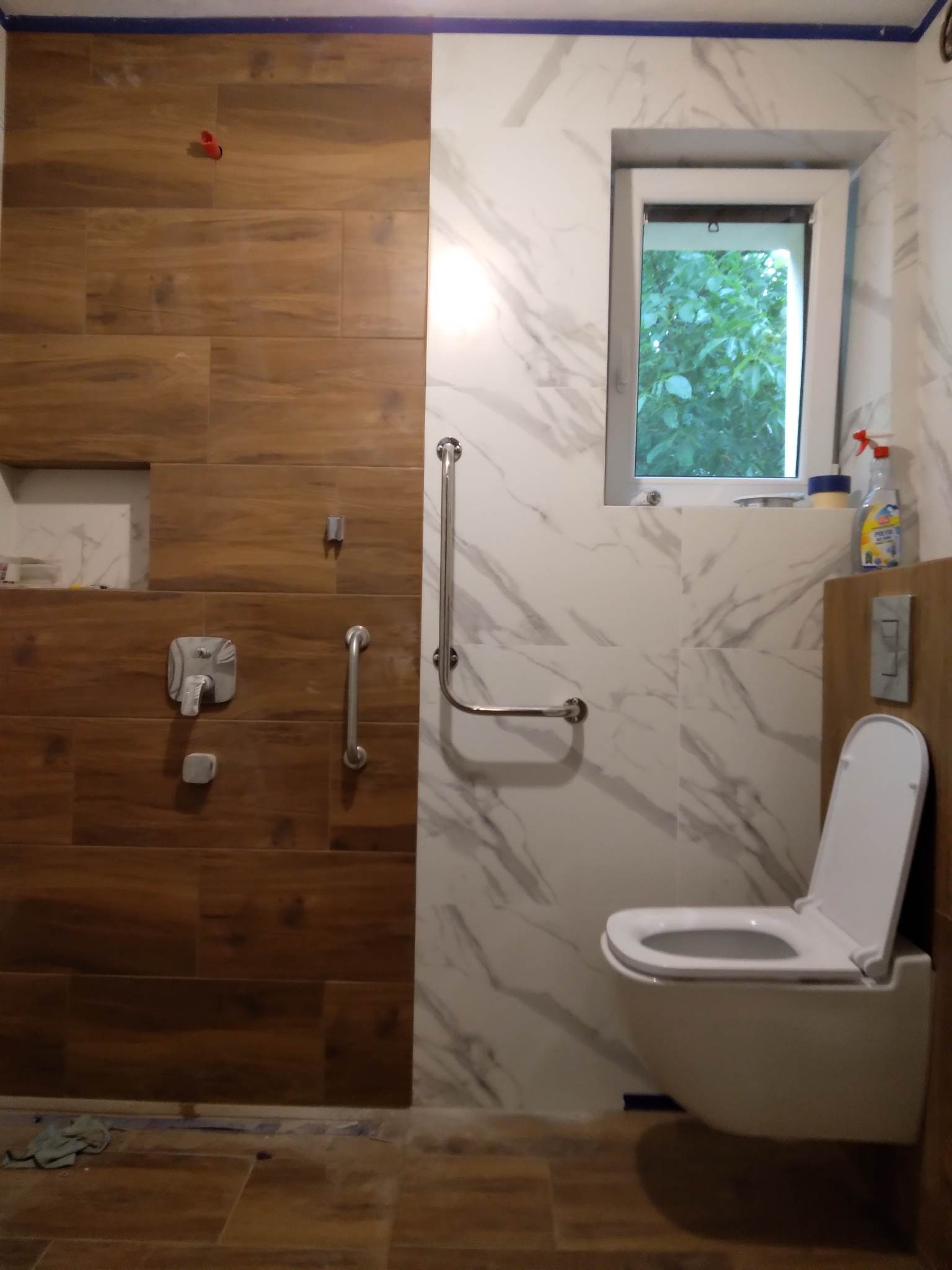 Nowoczesna łazienka w trakcie wykańczania: brązowe płytki imitujące drewno, białe marmurowe płytki, wisząca toaleta i uchwyt dla niepełnosprawnych. Widok na okno.