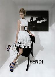 Modelka w białej sukience z ażurowym dołem i szerokim paskiem, trzymająca białą torebkę, pozująca na czarnym krześle na tle czarno-białego obrazu, z widocznym logo Fendi.