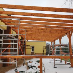 Such bud - Drewniana konstrukcja z belek, prawdopodobnie zadaszenie tarasu, w trakcie budowy. Widoczne rusztowania i śnieg na ziemi. Jasne drewno kontrastuje z żółtą elewacją budynku.