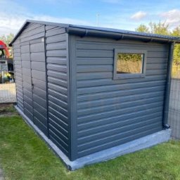 Such bud - Nowoczesna, grafitowa wiata narzędziowa z oknem, wykonana z paneli, z rynną i rurą spustową, na betonowej podmurówce, otoczona zielonym trawnikiem i ogrodzeniem.