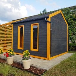 Such bud - Czarna, drewniana altana z żółtymi obramowaniami okien i dachu, otoczona zielenią i kwiatami w donicach. Nowoczesny design i staranne wykonanie.