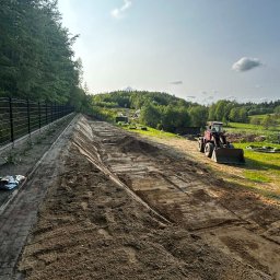 Mateusz Klein KLEIN Prace Ziemne - Równanie terenu pod przyszły ogród z użyciem traktora, widok z góry na przygotowany teren, obok ogrodzenie i zieleń. Prace ziemne w toku.