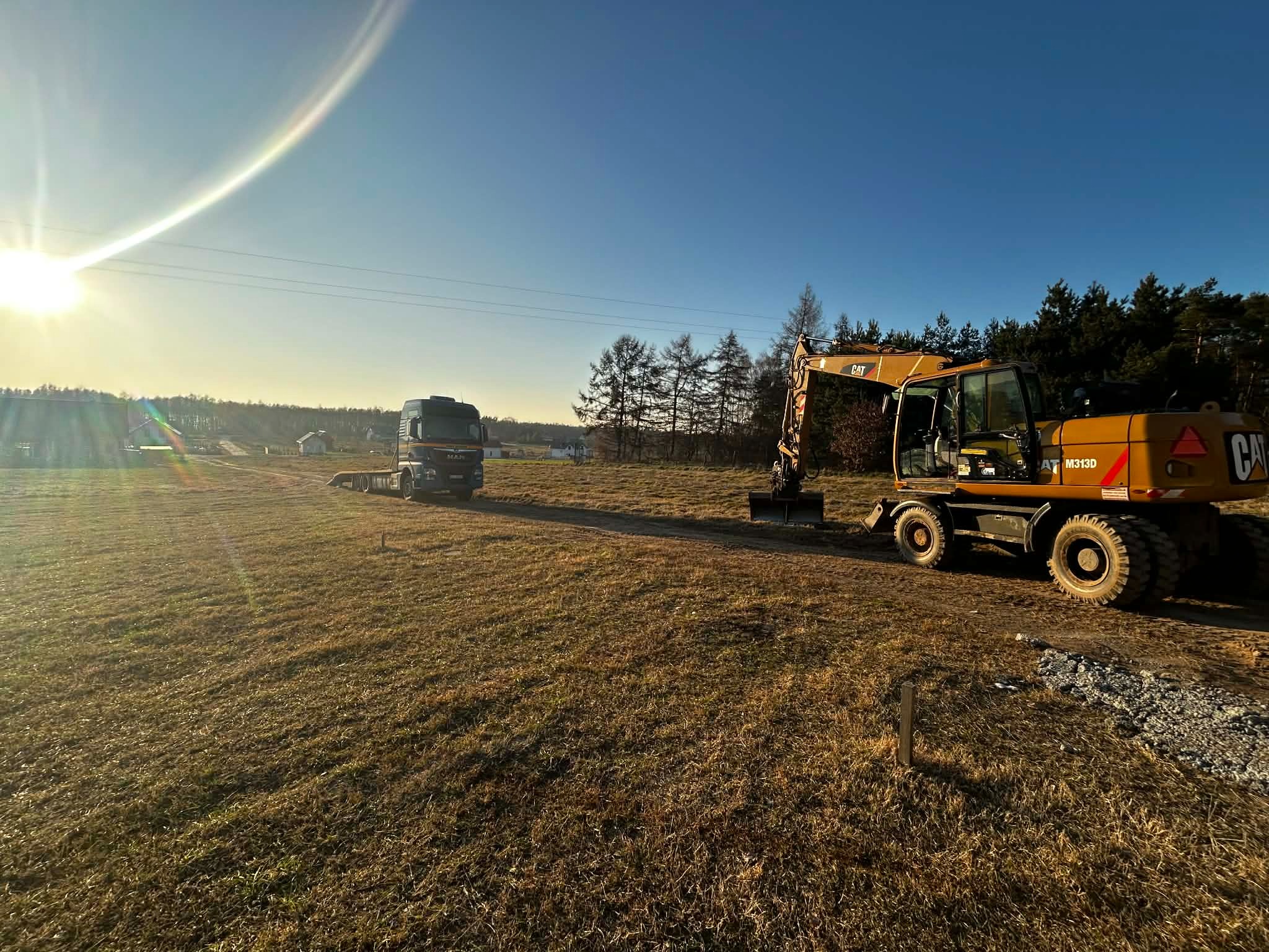 Żółta koparka CAT M313D i ciężarówka MAN na polu w słoneczny dzień. Widoczne drzewa w tle. Przygotowanie terenu pod budowę.