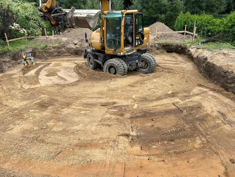 Żółta koparka Caterpillar w okrągłym wykopie, widoczne ślady opon i ubijarka w tle. Wykop otoczony zielenią, częściowo zabezpieczony drewnianą konstrukcją.