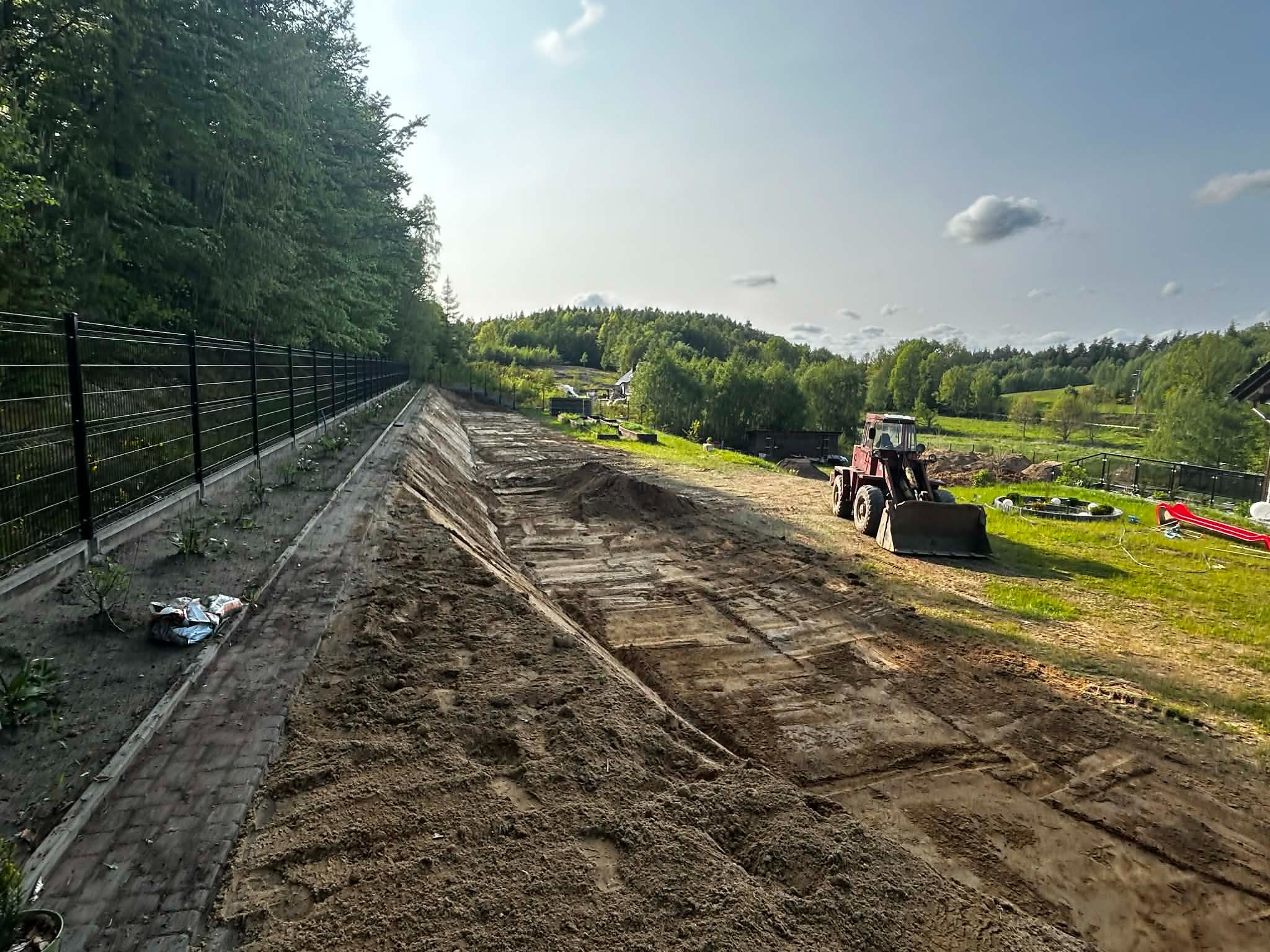 Równanie terenu pod przyszły ogród z użyciem traktora, widok z góry na przygotowany teren, obok ogrodzenie i zieleń. Prace ziemne w toku.