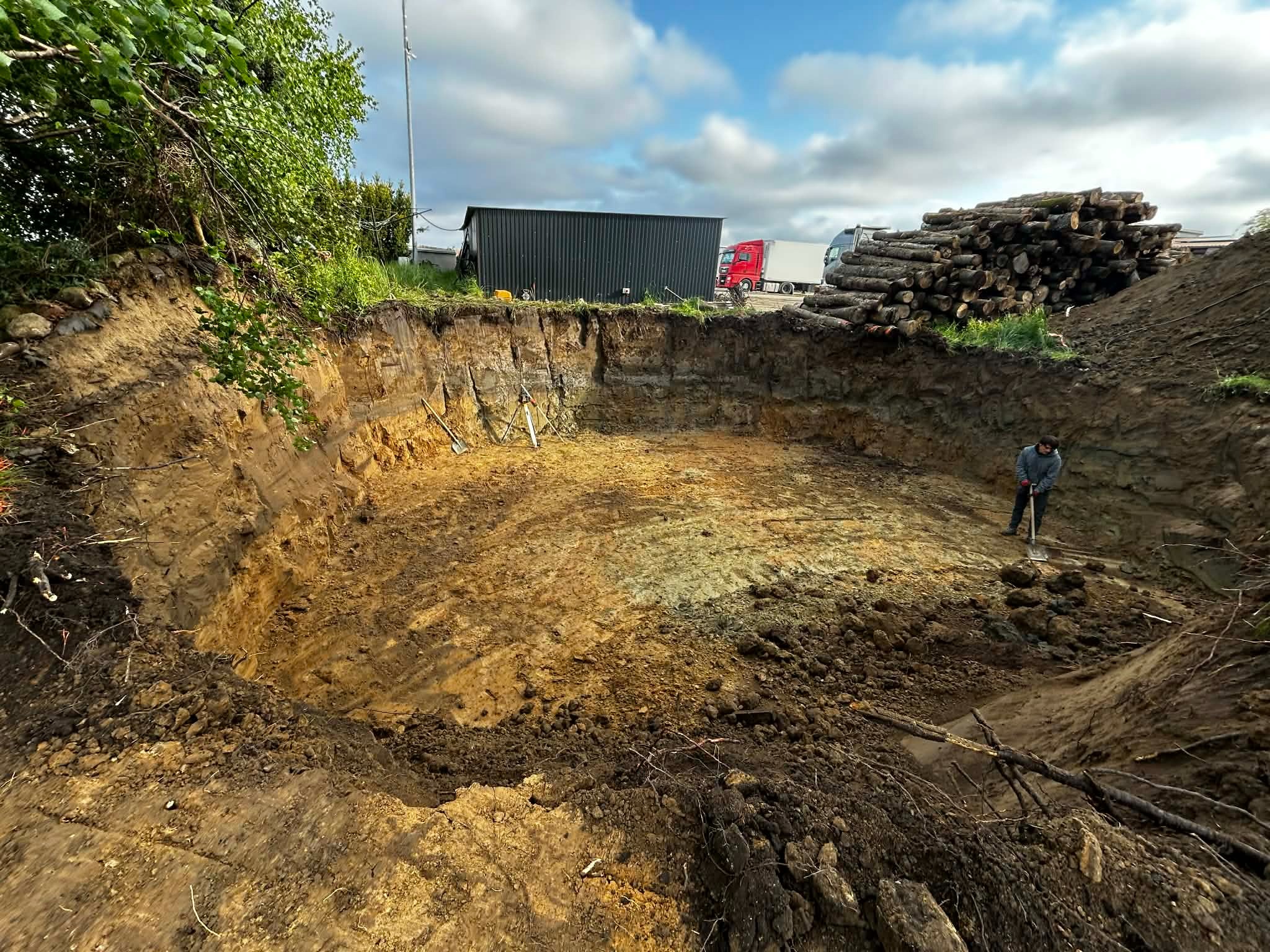 Głęboki, okrągły wykop w ziemi, z widoczną warstwową strukturą gleby. W tle sterta ściętych pni drzew, budynek gospodarczy i ciężarówki. Mężczyzna z łopatą w dole wykopu.