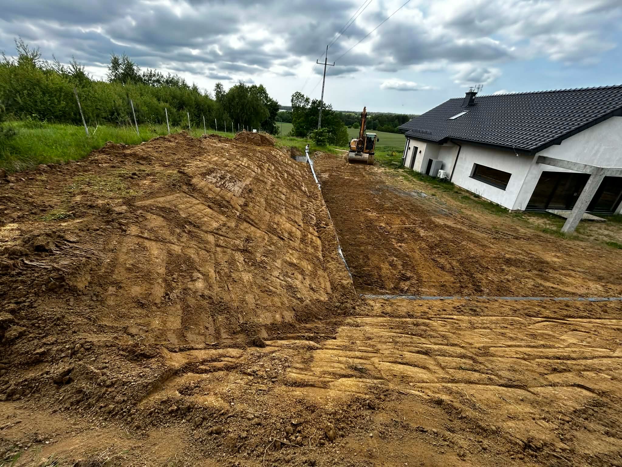 Niwelacja terenu pod budowę domu jednorodzinnego. Widoczny wykop, ziemia z wyraźnymi śladami opon i koparka w tle. Prace ziemne w toku, pochmurne niebo.