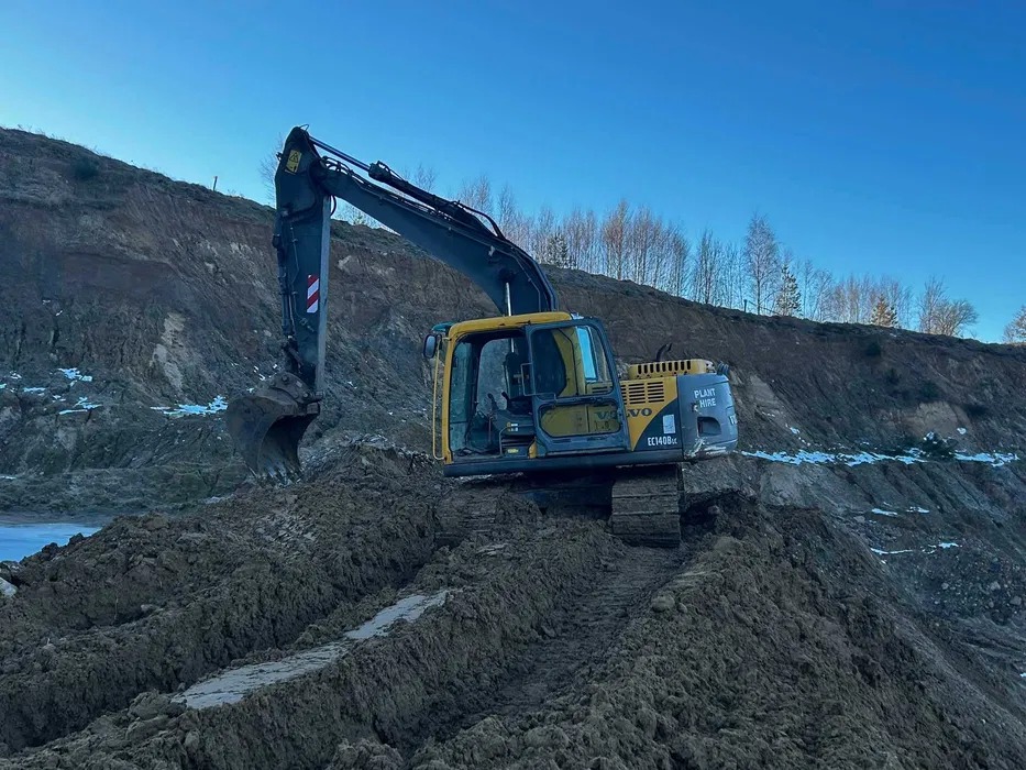 Żółto-szara koparka Volvo EC140B LC na nierównym, błotnistym terenie z wykopem, na tle wzgórza i błękitnego nieba. Widoczne ślady gąsienic.
