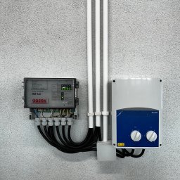 Szara ściana z zamontowanym modułem alarmowym Gazex MD-4.Z, białą skrzynką SC2A1 z niebieskim panelem i trzema białymi rurkami z kablami. Instalacja zabezpieczeń gazowych.