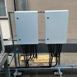 Dwie białe skrzynki elektryczne z czarnymi kablami, zamontowane na metalowej konstrukcji na zewnątrz budynku. Widoczne elementy instalacji i fragment elewacji.