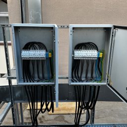 Dwa otwarte panele elektryczne z widocznymi przewodami i bezpiecznikami, zamontowane na metalowej konstrukcji na zewnątrz budynku. Widoczne kable w kolorach czarnym, niebieskim i żółto-zielonym.