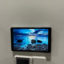 Panel sterowania inteligentnego domu z ekranem dotykowym, zamontowany na ścianie, obok termostatu. Widoczny interfejs użytkownika z informacjami o pogodzie i godzinie.