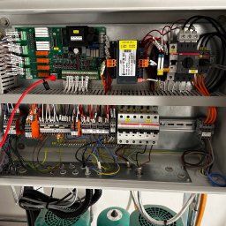 MK Electric System Marcin Kobrzyński - Otwarta szafa sterownicza z widocznymi elementami elektrycznymi: płytki drukowane, transformator, bezpieczniki, styczniki ABB i liczne przewody. Widoczne zielone silniki.