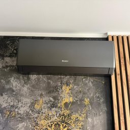 KLIMA-BROTHERS PIOTR ŚLIWKA - Szary klimatyzator marki Gree na ścianie z dekoracyjną fakturą w odcieniach szarości i złota, obok drewnianej ażurowej ścianki działowej z pionowych listew.