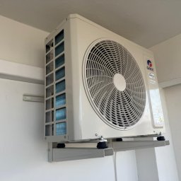 KLIMA-BROTHERS PIOTR ŚLIWKA - Zamontowana jednostka zewnętrzna klimatyzacji marki Gree na białej ścianie. Widoczne mocowania i fragment ściany z oknem. Ujęcie z dołu, lekko z lewej strony.