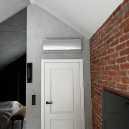 NexKlima Sebastian Niemczyk - Klimatyzator ścienny w nowoczesnym wnętrzu z ceglaną ścianą i białymi drzwiami. Widoczny fragment sypialni z łóżkiem. Szara ściana i skośny sufit.