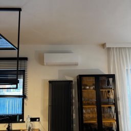 NexKlima Sebastian Niemczyk - Biały klimatyzator ścienny w nowoczesnym wnętrzu z czarnymi akcentami: grzejnik, okno z żaluzjami i witryna. Minimalistyczny design, jasne ściany.