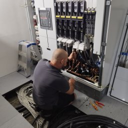 Mariusz Socha - Elektryk w szarej koszulce, w trakcie pracy nad rozbudowaną instalacją elektryczną w szafie sterowniczej. Widoczne kable, narzędzia i drabina.