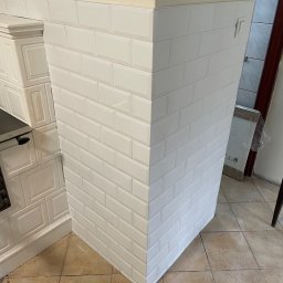 Perfect Wall - Wykończony narożnik z białych, prostokątnych płytek, ułożonych w cegiełkę. Widoczny fragment pieca kaflowego i beżowa ściana. Podłoga wyłożona płytkami w odcieniach beżu.