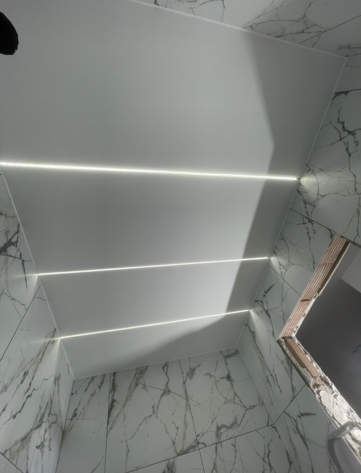 Nowoczesna łazienka z podwieszanym sufitem i liniowym oświetleniem LED, marmurowe płytki na ścianach, widok z dołu, surowy otwór drzwiowy.