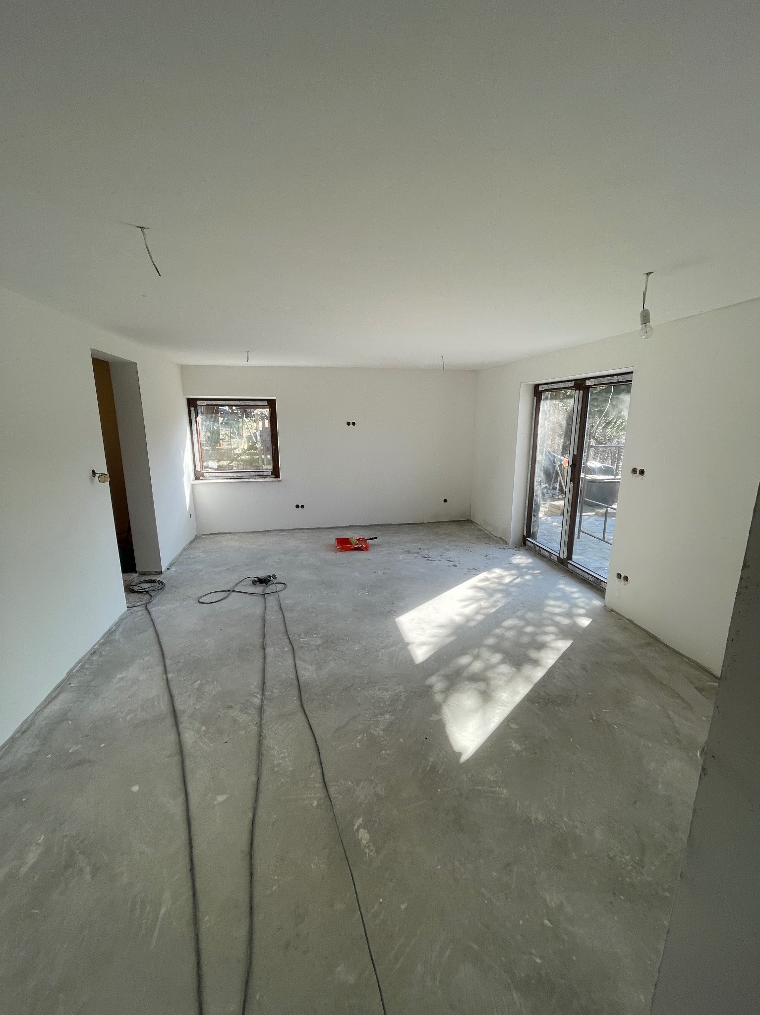 Wnętrze po remoncie: białe ściany, szara podłoga, widoczne kable i narzędzia malarskie. Duże okno z widokiem na zewnątrz. Przygotowanie do dalszych prac wykończeniowych.