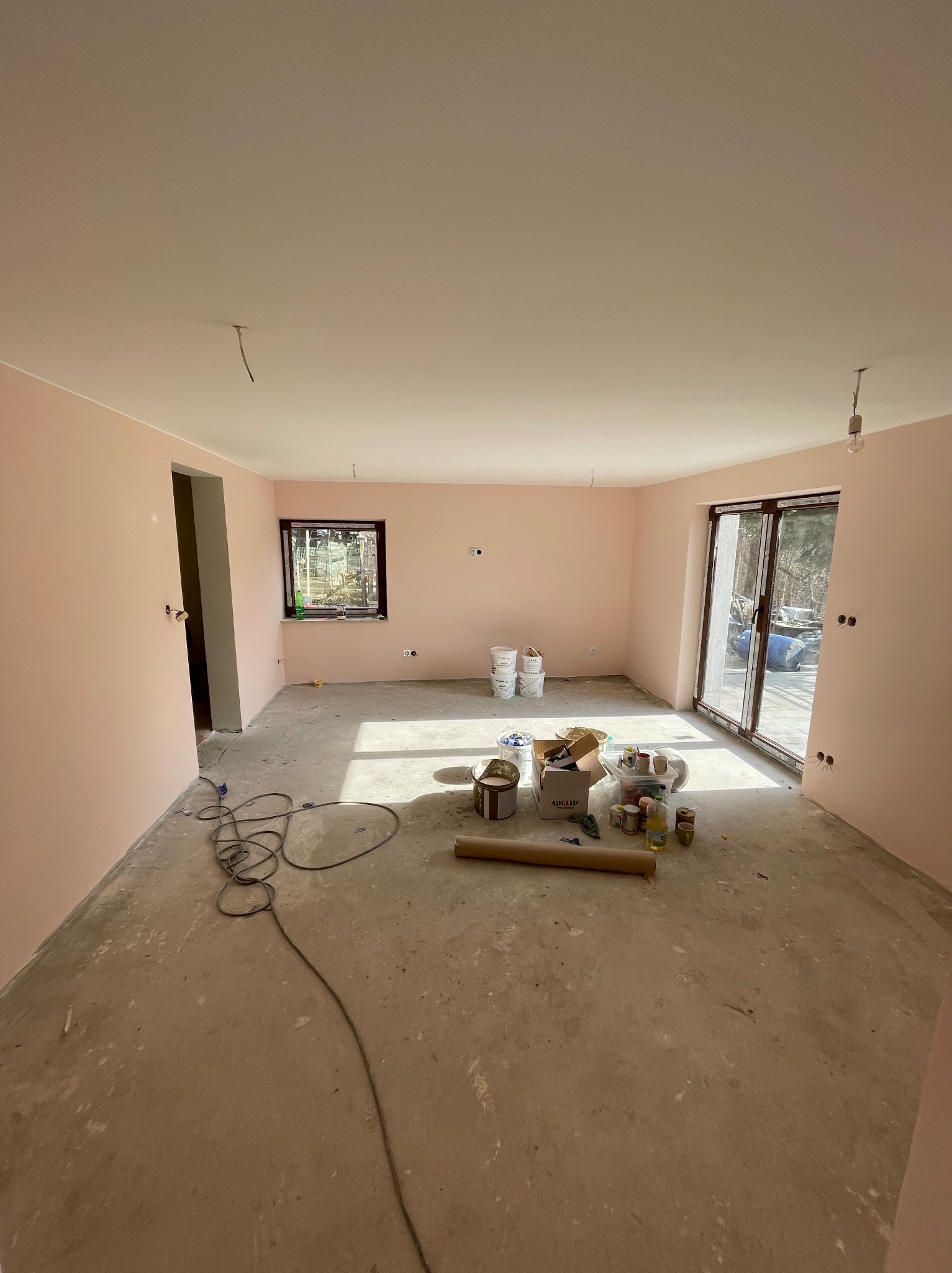 Pomieszczenie po malowaniu ścian na delikatny róż, widoczne puszki z farbą, wałki i kabel na podłodze. Okno i drzwi balkonowe w tle. Etap prac wykończeniowych.