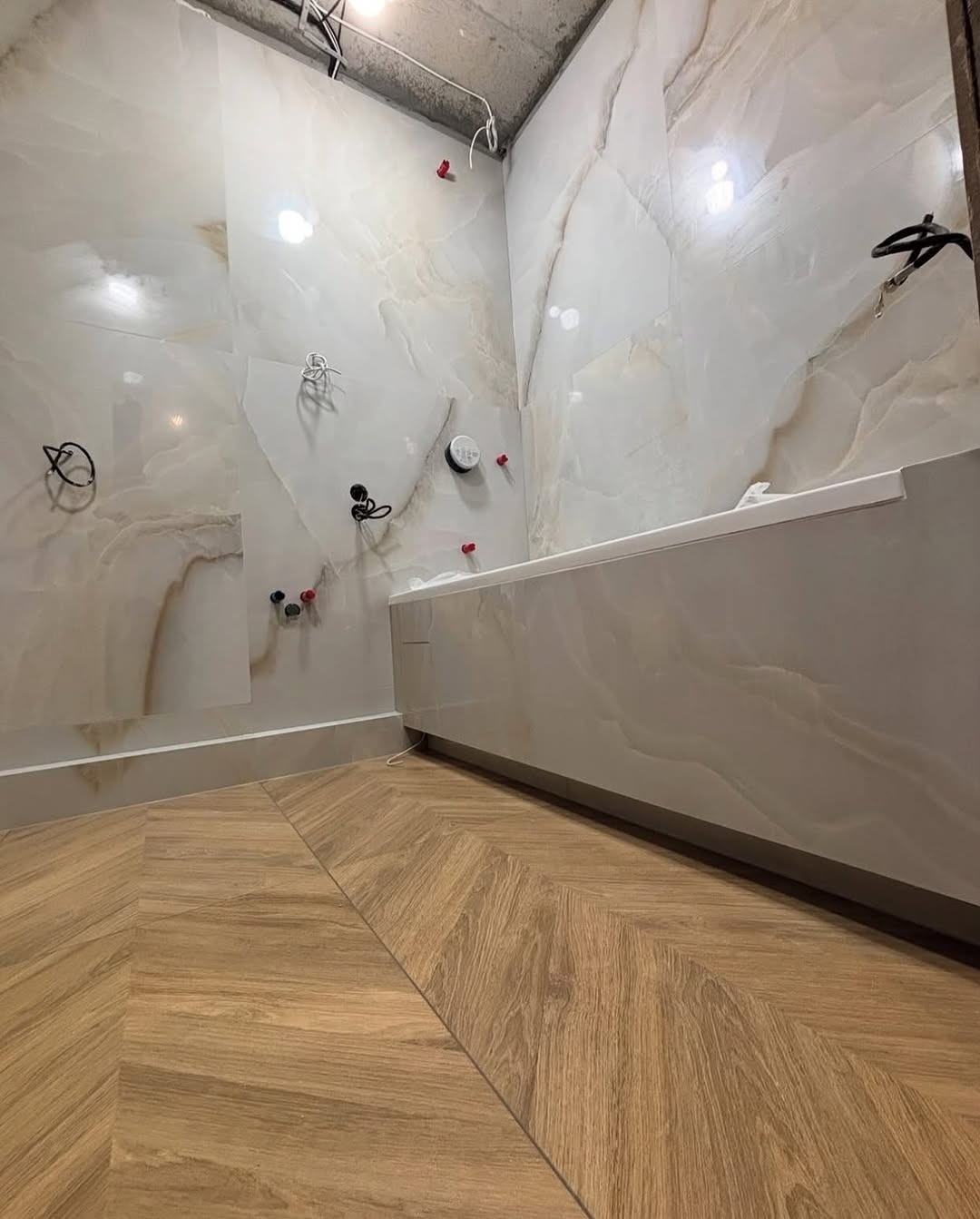 Nowoczesna łazienka w trakcie wykańczania: marmurowe płytki ścienne, drewnopodobna podłoga ułożona w jodełkę, widoczne instalacje przed montażem armatury.