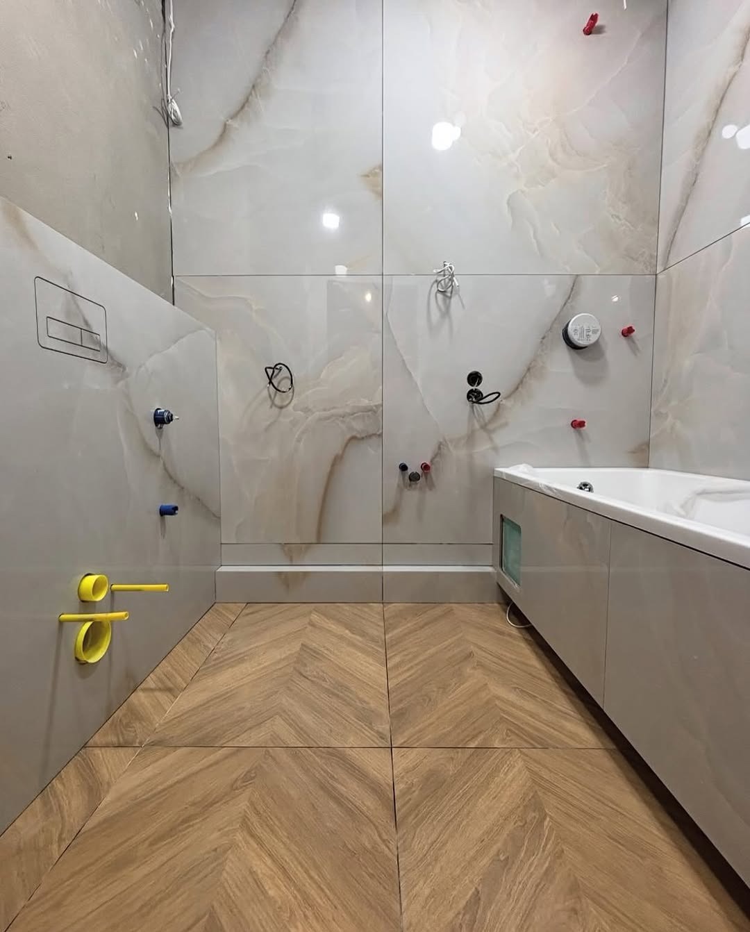 Nowoczesna łazienka w Warszawie z beżowymi płytkami imitującymi marmur na ścianach i drewnopodobną podłogą ułożoną w jodełkę, z widoczną wanną i instalacją.