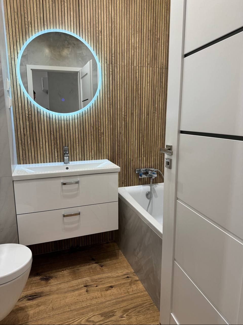 Nowoczesna łazienka z drewnianą ścianą, okrągłym lustrem z podświetleniem LED, białą szafką pod umywalką i wanną. Drewniana podłoga dodaje ciepła wnętrzu.