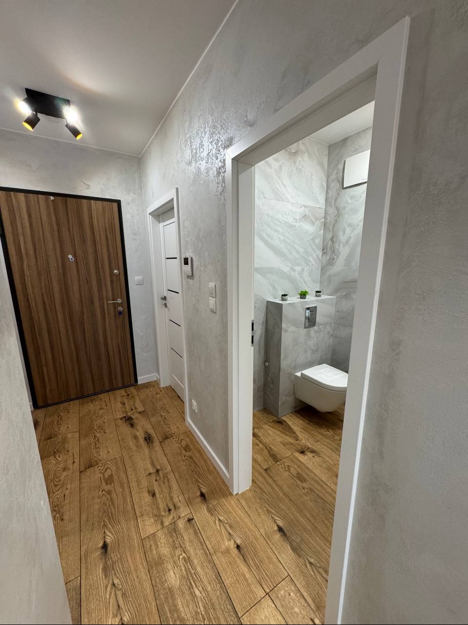 Wykończony korytarz z drewnianą podłogą i wejściem do łazienki z marmurowymi płytkami. Widoczne drzwi wejściowe i wewnętrzne. Ściany pokryte dekoracyjnym tynkiem.