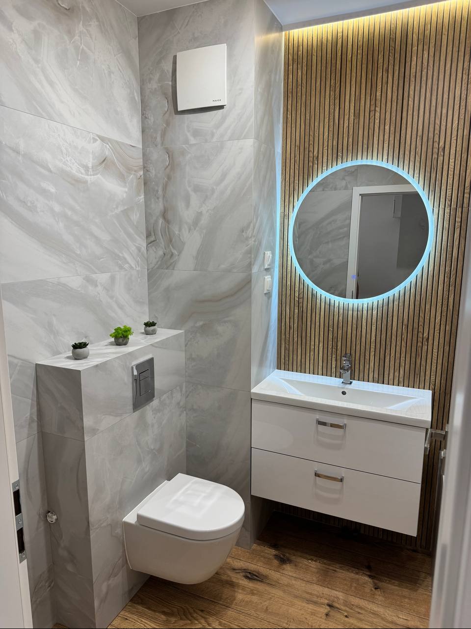 Nowoczesna łazienka z podwieszaną toaletą, umywalką z szafką i okrągłym lustrem z podświetleniem LED. Ściany wyłożone płytkami imitującymi marmur, drewniana okładzina.