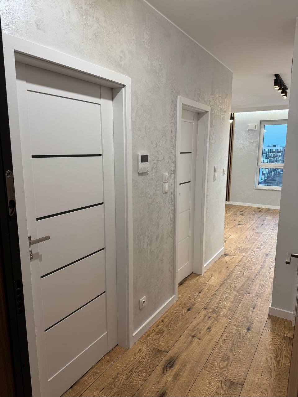 Nowoczesny korytarz po remoncie: drewniana podłoga, ściany z efektem dekoracyjnym, białe drzwi z czarnymi akcentami. Widok na dalszą część mieszkania.