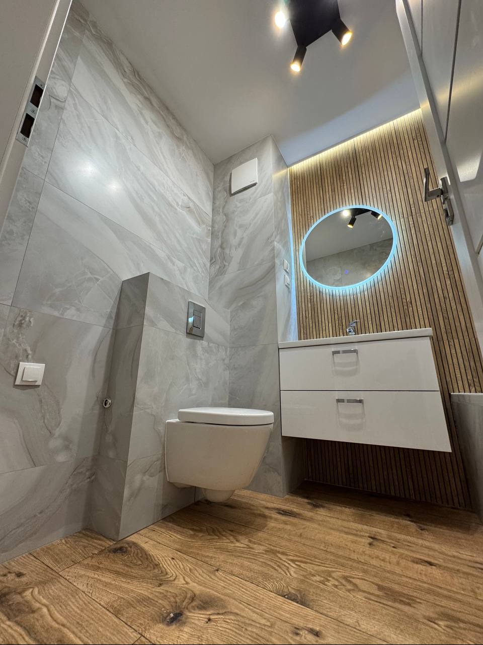 Nowoczesna łazienka z marmurowymi płytkami, drewnianą ścianą z podświetleniem LED za okrągłym lustrem i wiszącą szafką z umywalką. Drewniana podłoga.
