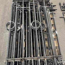 HUSSEG SPÓŁKA Z OGRANICZONĄ ODPOWIEDZIALNOŚCIĄ - Stos elementów kutych: fragmenty bramy z dekoracyjnymi detalami, w tym szpice i okręgi, ułożone na palecie w warsztacie. Surowy metal, gotowe do montażu.