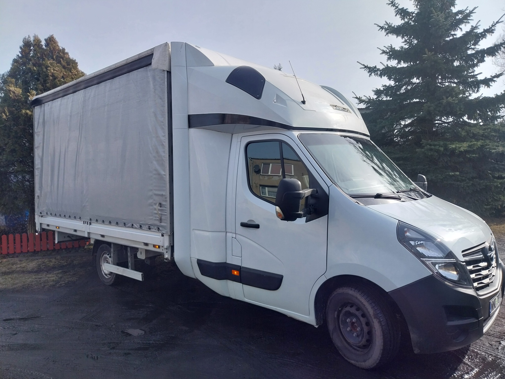 Biały bus z plandeką na tle zieleni, widok z boku. Charakterystyczna zabudowa kabiny kierowcy. Pojazd zaparkowany na utwardzonym terenie.