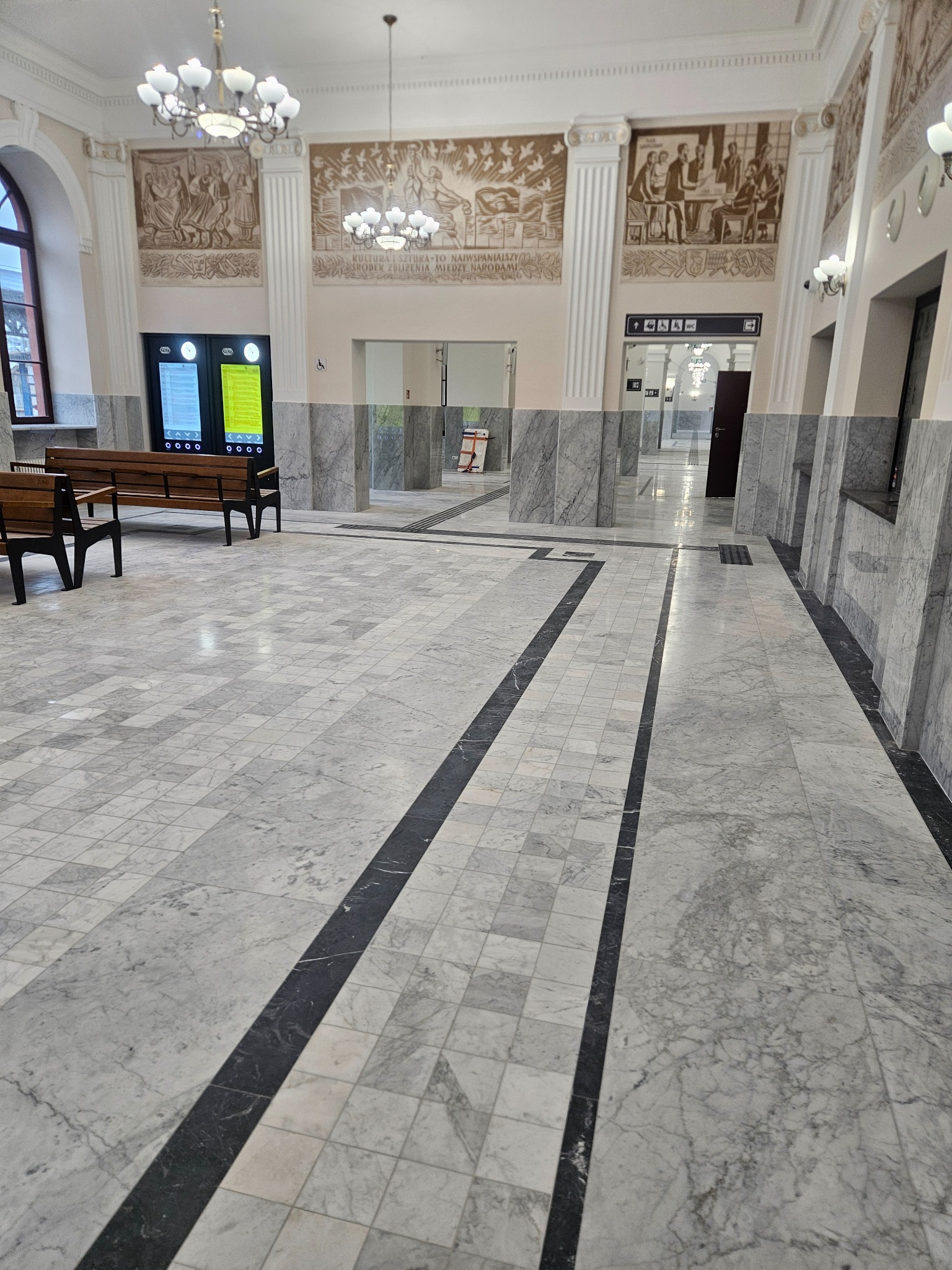 Przestronne wnętrze z marmurową posadzką w szaro-białe kwadraty, ławkami i dekoracyjnymi malowidłami na ścianach. Widoczne kolumny i oświetlenie.