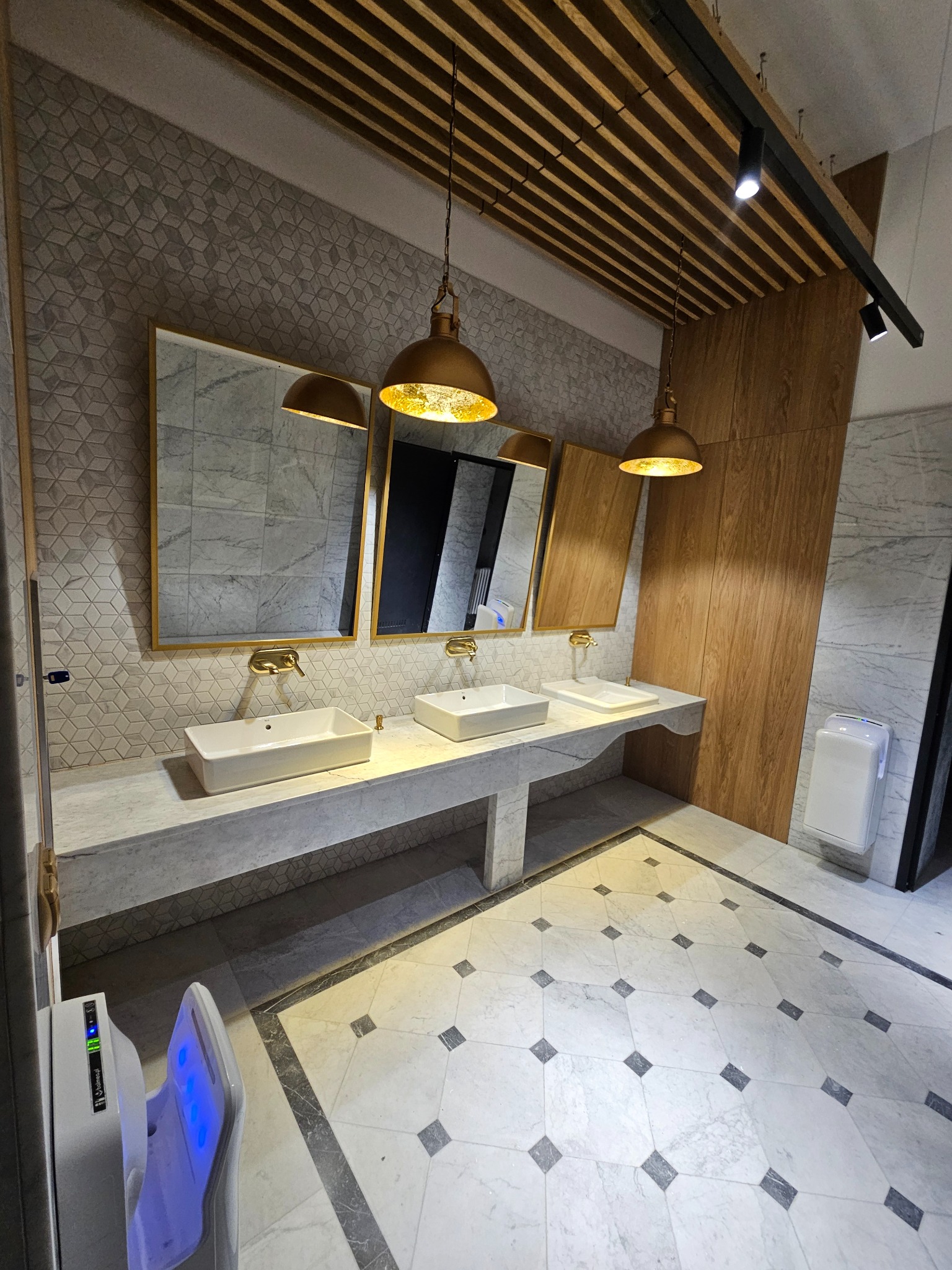 Elegancka łazienka publiczna z trzema umywalkami, lustrami w złotych ramach i marmurową podłogą w geometryczny wzór. Oświetlenie punktowe i drewniane panele na suficie.