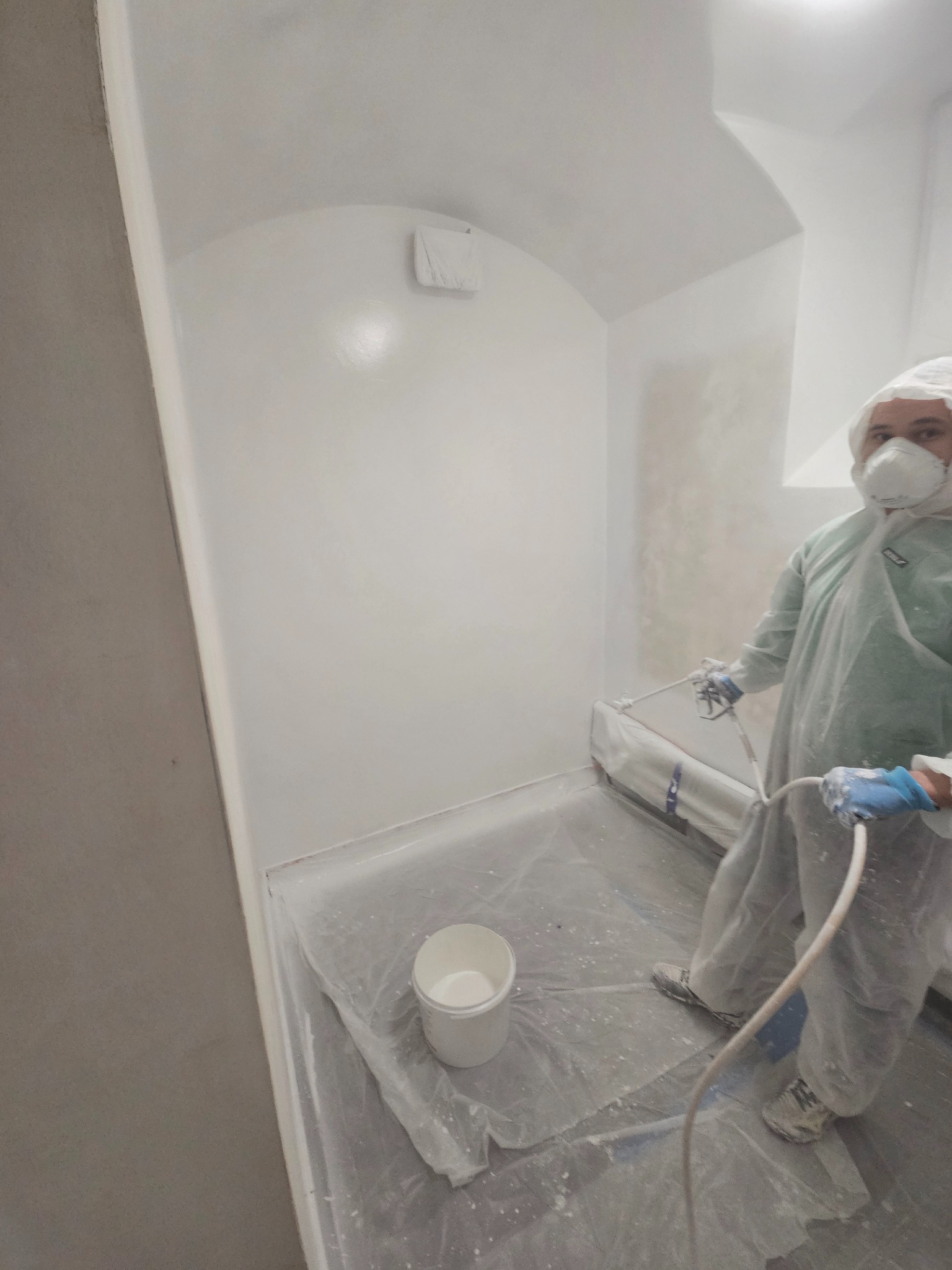 Pracownik w kombinezonie ochronnym maluje białą farbą wnętrze pomieszczenia za pomocą pistoletu natryskowego. Podłoga zabezpieczona folią, widoczne wiadro z farbą.