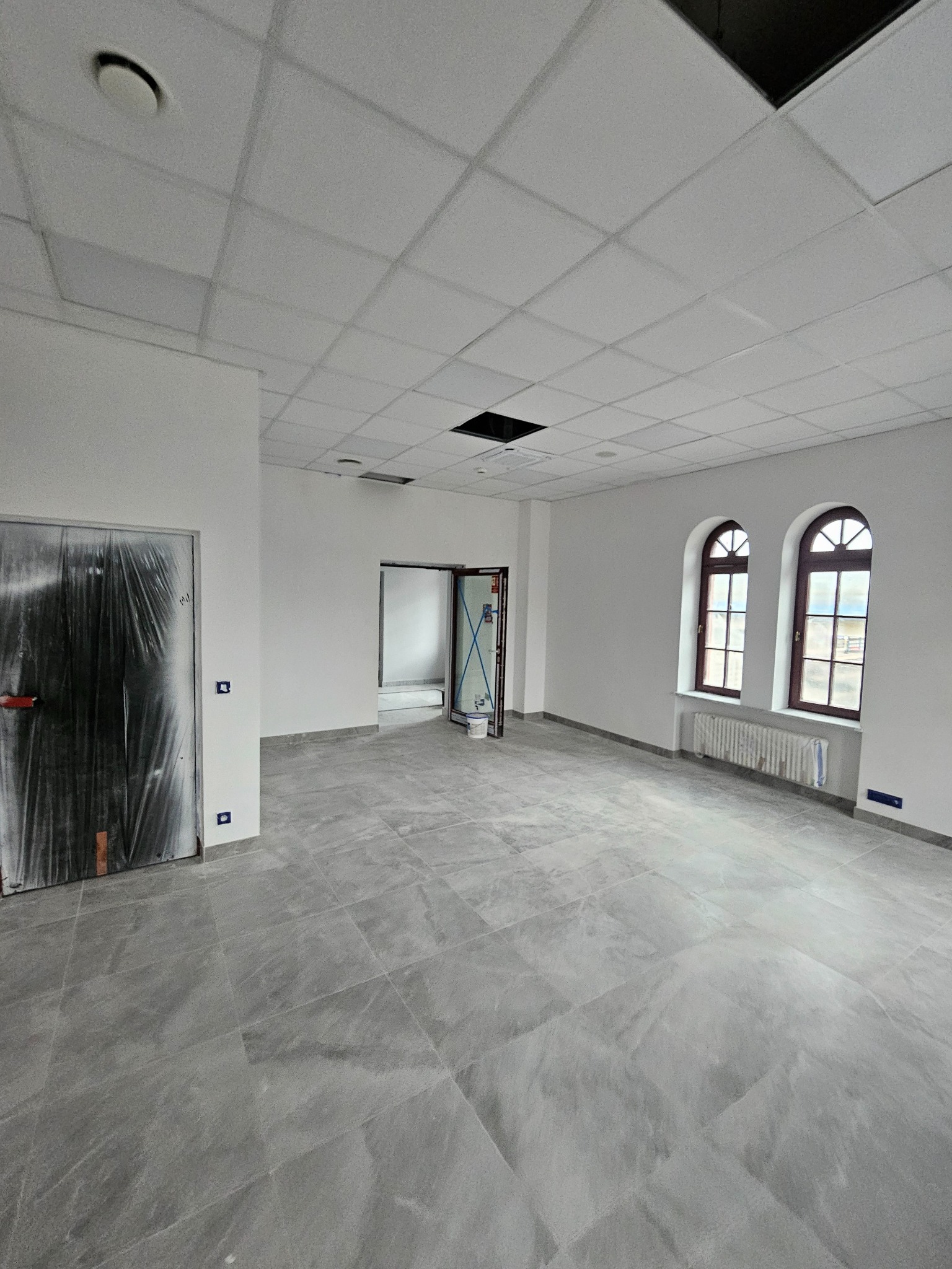 Wnętrze biura po remoncie: szara podłoga z płytek, białe ściany, okna z łukami. Drzwi zabezpieczone folią malarską. Widok na sąsiednie pomieszczenie.
