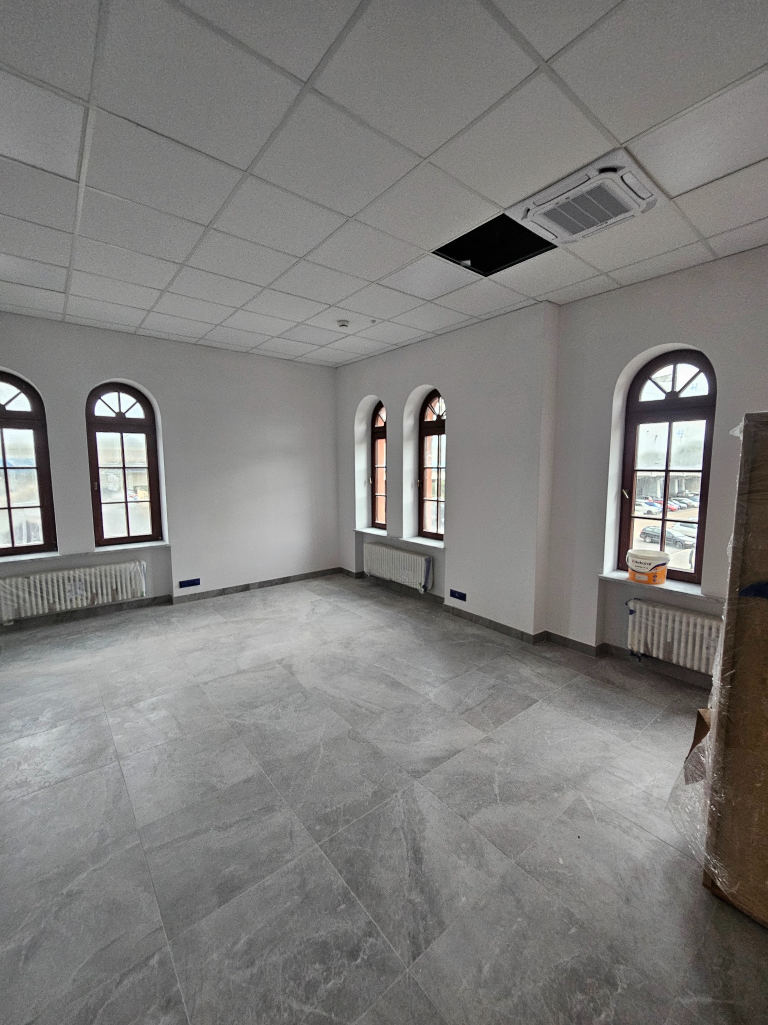 Puste, jasne biuro po remoncie z szarą podłogą z płytek, białymi ścianami, brązowymi oknami łukowymi i grzejnikami. Widoczny klimatyzator i wiadro z farbą.