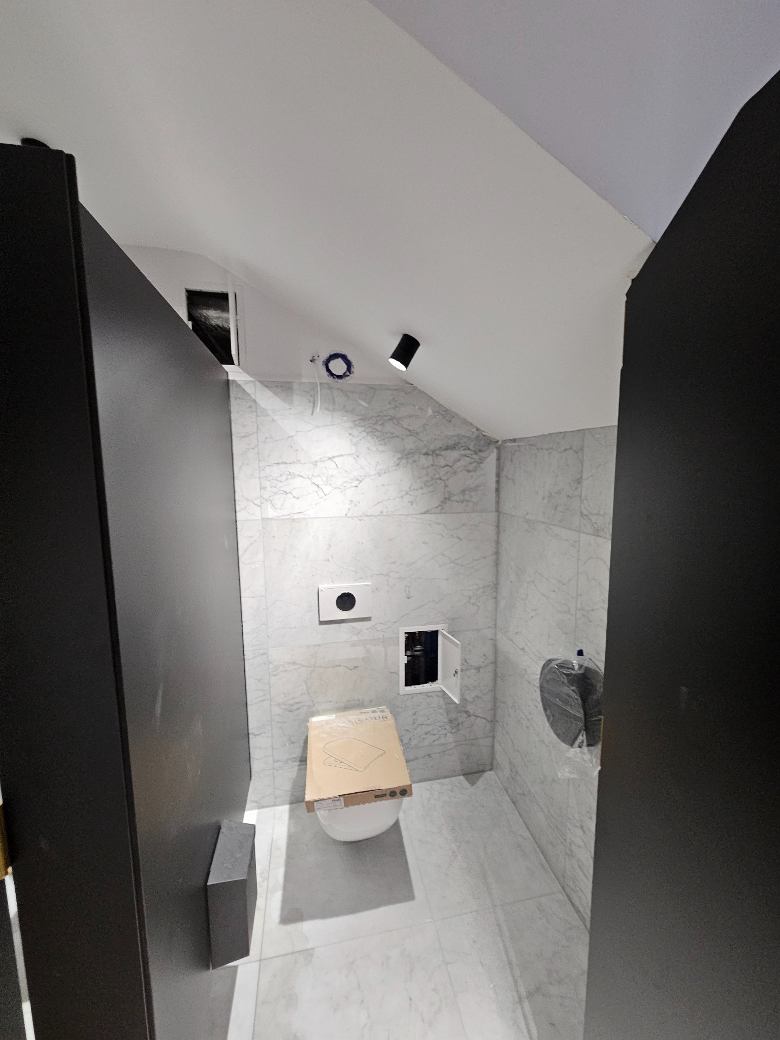 Nowoczesna łazienka w trakcie wykańczania: białe marmurowe płytki, czarne drzwi, podwieszana toaleta z kartonem na sedesie. Oświetlenie punktowe.