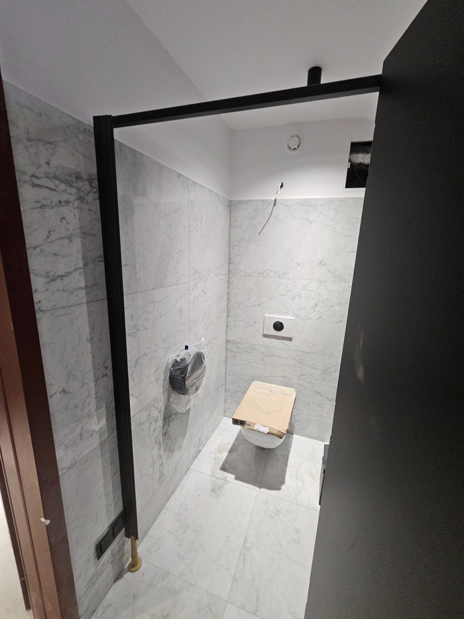 Wnętrze łazienki w trakcie wykańczania: marmurowe płytki, czarna rama kabiny prysznicowej, częściowo zamontowana toaleta z kartonem na sedesie. Widoczne elementy instalacji.