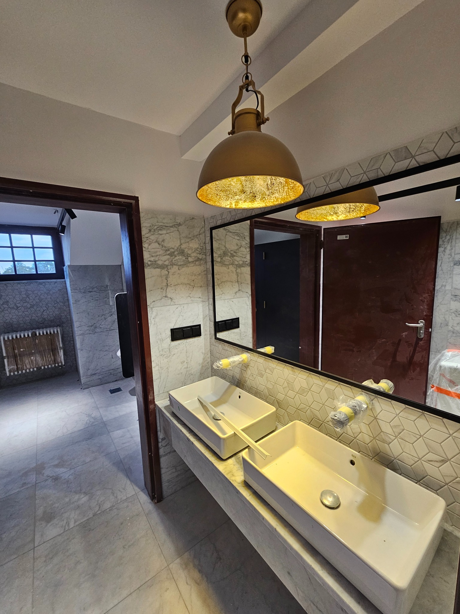 Wykończona łazienka z marmurowymi płytkami, podwójną umywalką i złotą lampą wiszącą. Widoczne lustro i drzwi do innego pomieszczenia. Nowoczesny design.