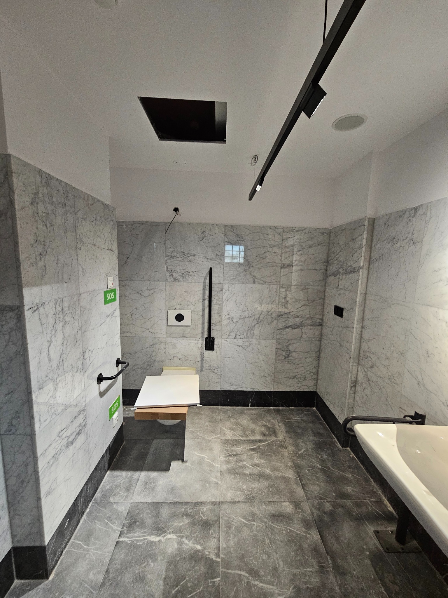 Wykończona łazienka w szarym marmurze z udogodnieniami dla osób niepełnosprawnych: poręcze, siedzisko pod prysznicem, przycisk SOS. Nowoczesne oświetlenie.