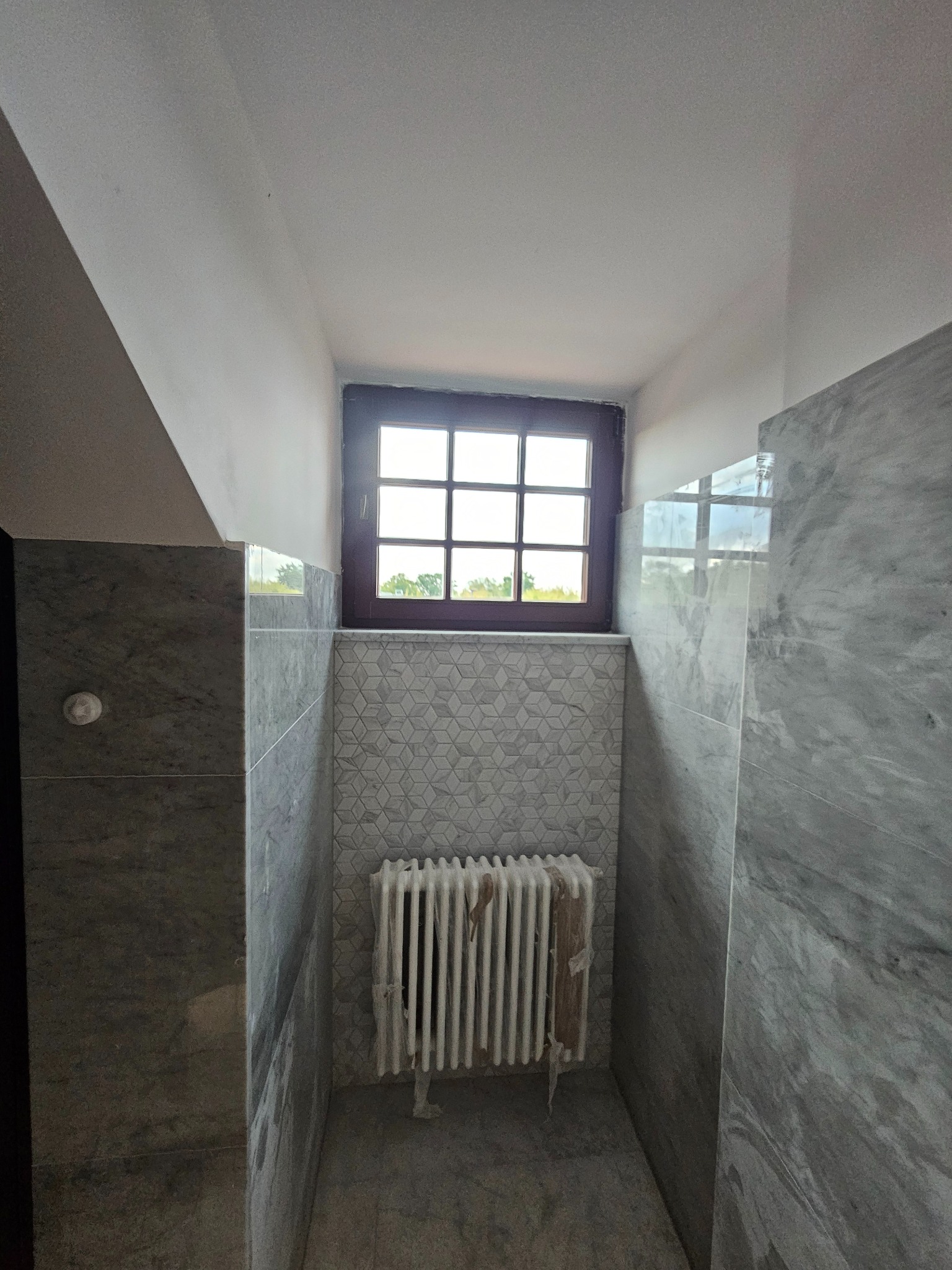 Wykończona łazienka z marmurowymi płytkami, grzejnikiem osłoniętym folią i oknem z widokiem na zieleń. Geometryczne płytki nad grzejnikiem. Nowoczesny design.