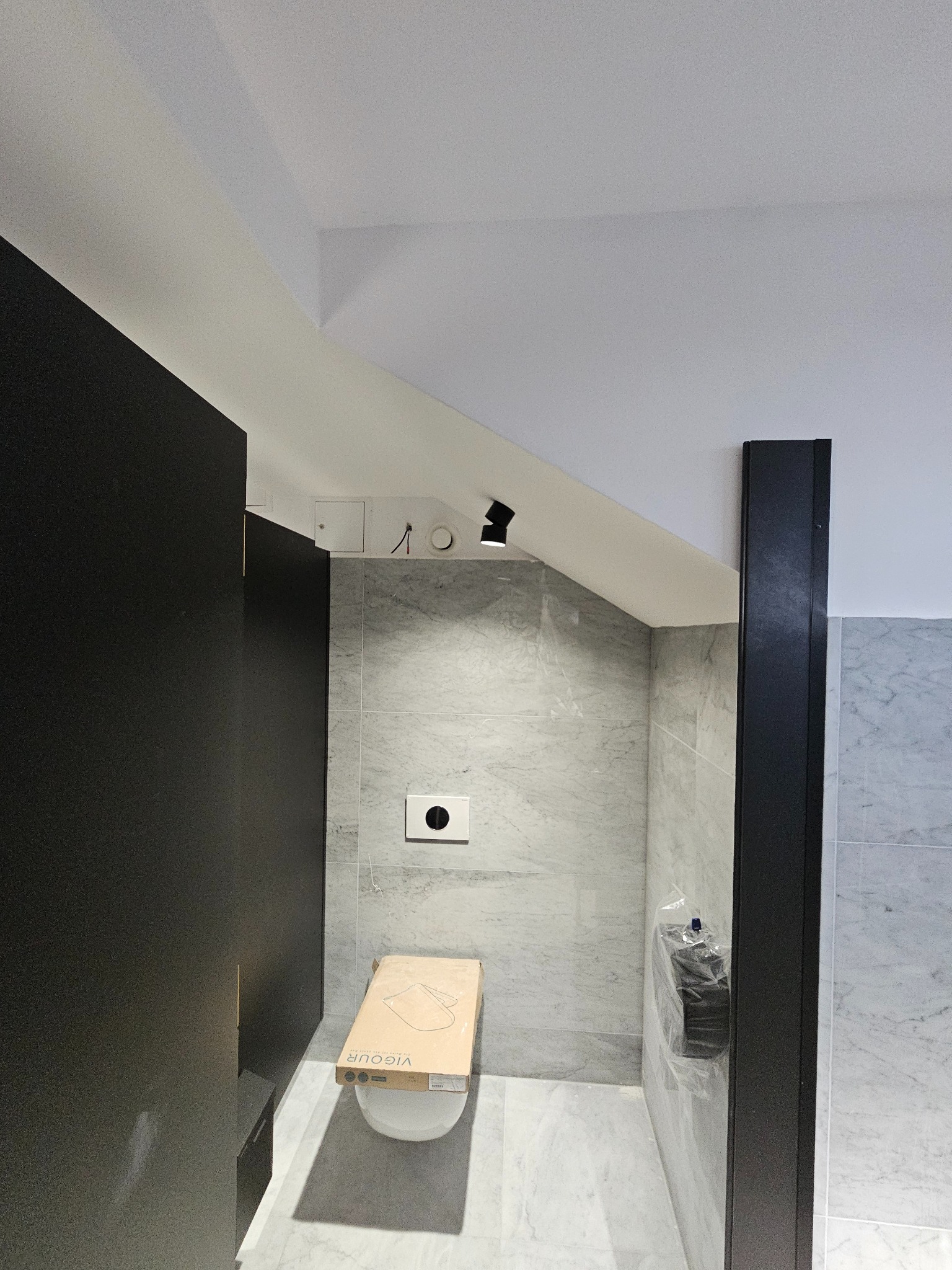 Nowoczesna łazienka w trakcie wykańczania: marmurowe płytki, czarne elementy, toaleta w kartonie. Minimalistyczny design, sufit ze skośnym załamaniem, oświetlenie punktowe.