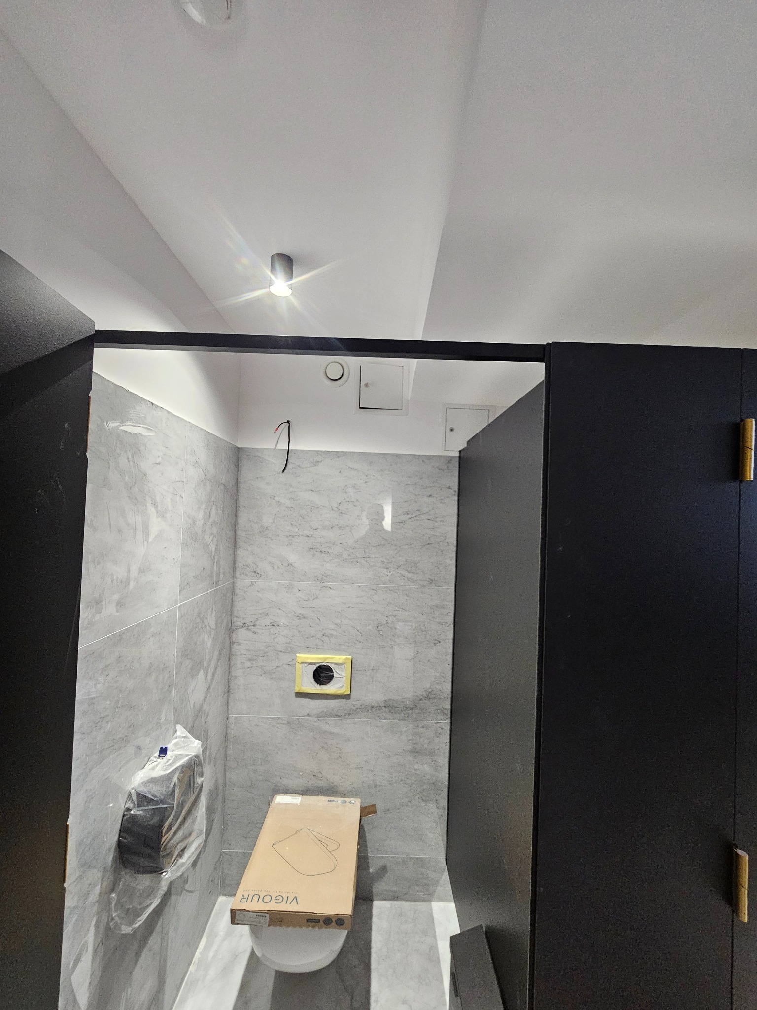 Wnętrze łazienki w trakcie wykańczania: szare płytki na ścianach, zabudowa meblowa w kolorze czarnym, częściowo zamontowana toaleta w kartonie.