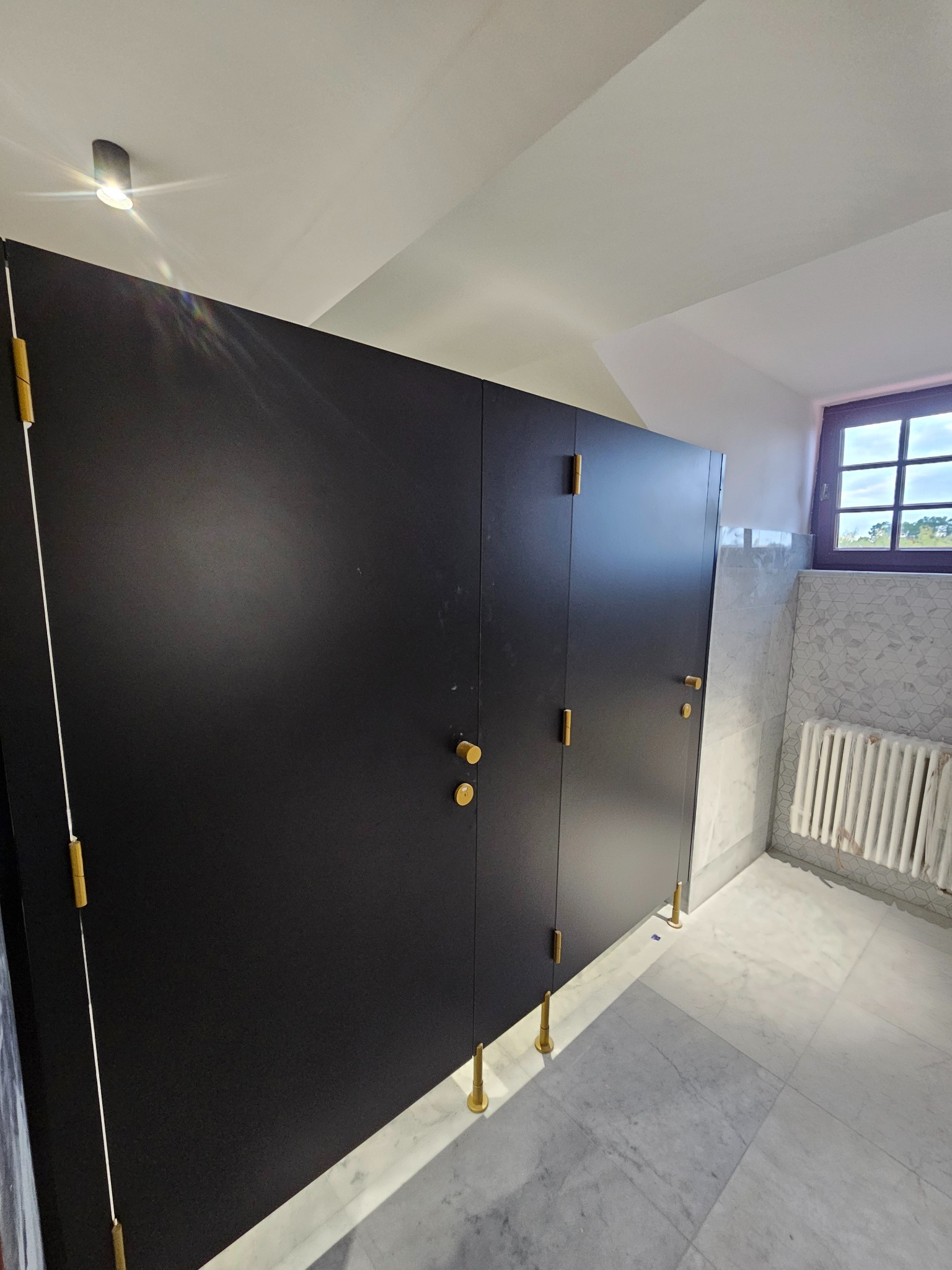 Czarne drzwi kabin WC z złotymi detalami w wykończonej łazience z marmurową podłogą i grzejnikiem pod oknem.